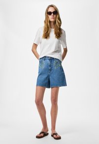 Femme portant des lunettes de soleil noires, un t-shirt blanc, un short en jean avec broderie florale et des sandales marron foncé, debout les mains derrière le dos.