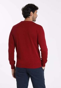 Pull en tricot rouge à manches longues, avec des poignets et un ourlet côtelés, doté d'un col rond classique et d'une texture lisse. Porté avec un pantalon bleu marine.
