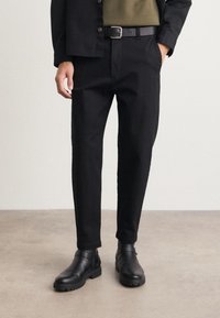 Black tapered trousers made of smooth fabric, featuring front pockets and a zipper fly, paired with black leather ankle boots.