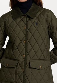 Vrouw in een donkergroene gewatteerde jas met corduroy kraag, drukknoopjes en voorzakken over een donkere turtleneck.