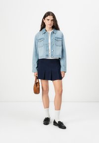 Lyseblå jeansjakke, hvit topp, marineblå plisserte skjørt, svarte loafer-sko og brun håndveske. Hvite sokker synlige. En enkel, uformell antrekk.