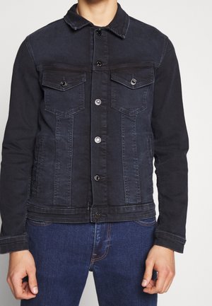 Mężczyzna w czarnej kurtce jeansowej na guziki z dwoma klapowanymi kieszeniami na piersi, połączony z niebieskimi dżinsami.