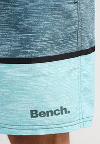 Plavkové šortky v prechode od tyrkysovej po modrú, s textúrovaným vzorom a čiernym akcentovým pruhom. Na spodnej strane je potlačené logo "Bench.".