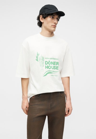 Νέος άνδρας που φοράει μαύρο καπέλο, λευκό μπλουζάκι oversized με πράσινο γράφημα "Döner House" και καφέ παντελόνι, στέκεται μπροστά σε απλό φόντο.