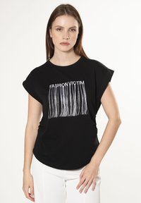 Camisa negra de manga corta con el texto blanco 'FASHION VICTIM' y un diseño con flecos. Material de algodón suave, corte holgado, escote redondo.