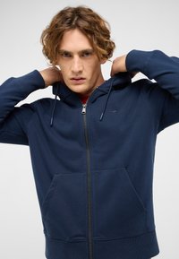 Navyblauer Kapuzenpullover mit Reißverschluss aus weichem Material, mit einer Fronttasche, Kordelzügen und gerippten Bündchen. Kleines Logo auf der Brust.