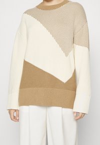 Žena v béžovém a bílém pleteném svetr s širokými manžetami a bílými kalhotami s vysokým pasem, stojící na jednotném pozadí.