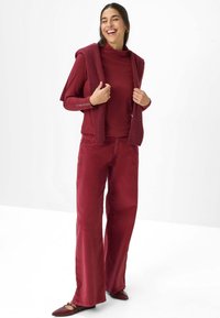 Burgunderfarbendes Outfit mit einem figurbetonten Rollkragenpullover und weiten Cordhosen. Eine passende Strickjacke ist über die Schultern drapiert. Glatte Textur.