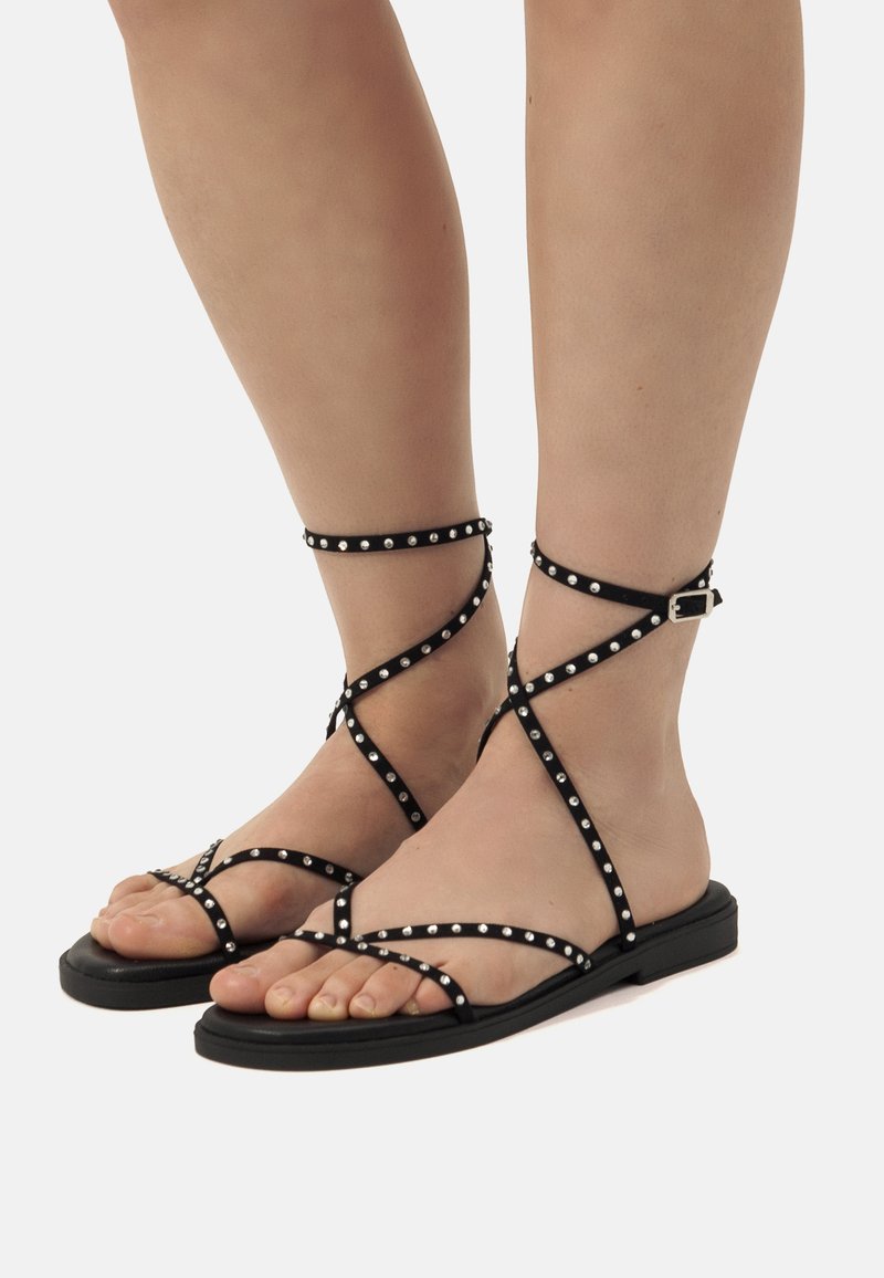 Las sandalias planas negras presentan tiras cruzadas adornadas con tachuelas metálicas, un diseño de punta abierta y un cierre con hebilla en el tobillo.