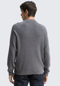 Szary sweter robiony na drutach z prążkowanymi detalami, okrągłym dekoltem i długimi rękawami. Materiał wydaje się miękki i teksturowany, noszony z ciemnymi dżinsami.