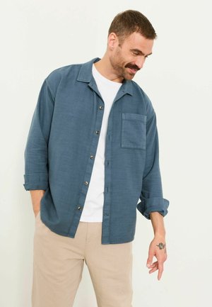 Mann mit kurzen Haaren und Schnurrbart, der ein blaues Hemd über einem weißen T-Shirt und beige Hose trägt, steht mit entspannten Händen.