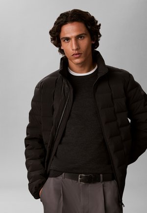 Jeune homme aux cheveux bouclés portant une doudoune noire, un pull noir, un pantalon gris et une ceinture noire, debout les mains dans les poches.