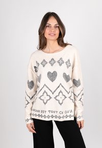Heller beiger Pullover mit grauen geometrischen Mustern, einschließlich Herzen und Rauten. Hat einen weit geschnittenen Halsausschnitt und lange Ärmel. Weiche Textur.
