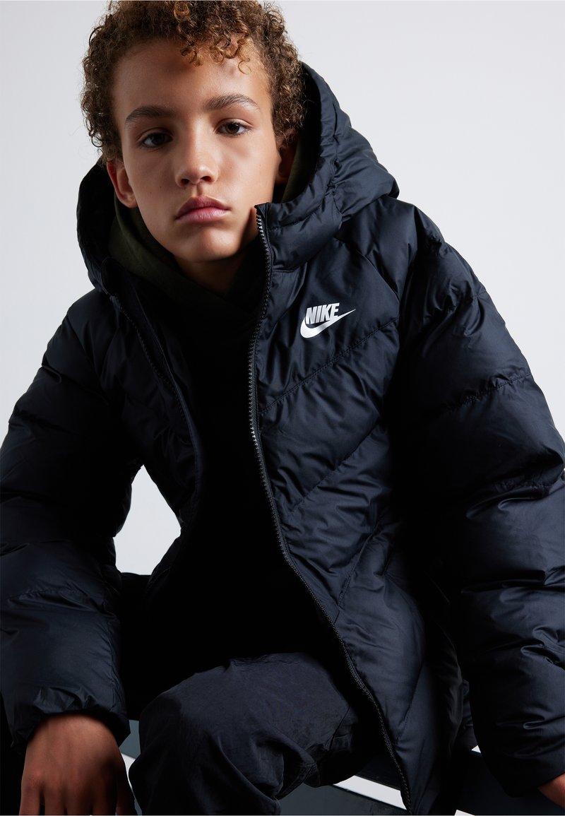 Czarna kurtka puchowa z kapturem, z pikowanym wzorem i białym logo Nike na piersi. Nosi się ją na ciemnozielonym hoodie.