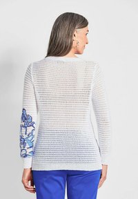 Weißer Strickpullover mit lockerem, offenem Design. Der linke Ärmel ist mit blauer Blumenstickerei verziert. Das Kleidungsstück hat einen Rundhalsausschnitt und einen gerippten Saum.