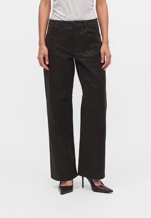 Personne portant un pantalon noir taille haute à jambes larges avec poches avant et des chaussures à talons hauts pointues et foncées, debout devant un fond uni.