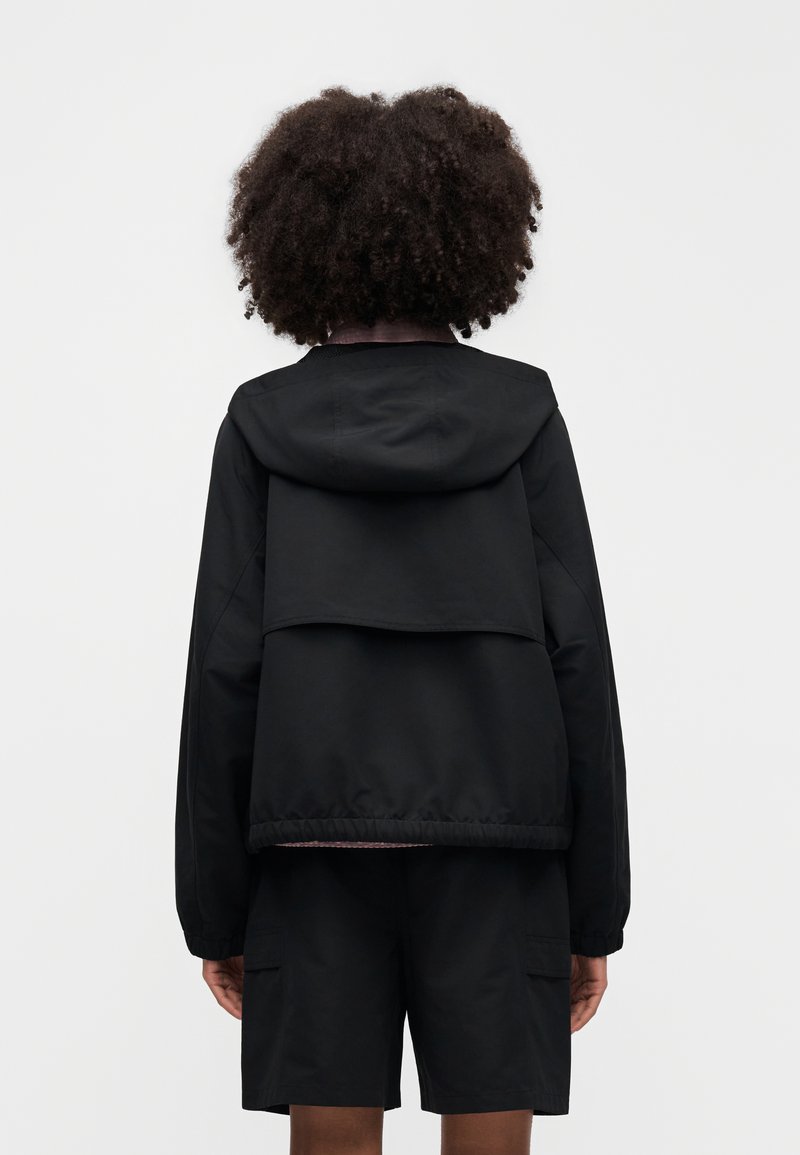 Giacca nera con cappuccio, con design posteriore a strati e polsini elasticizzati, abbinata a pantaloncini neri. Texture liscia, dettagli minimalisti.