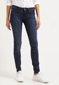 Jeans ajustados en mezclilla azul oscuro con una textura suave, que cuentan con bolsillos frontales y un cierre de botón, complementados con zapatillas blancas.