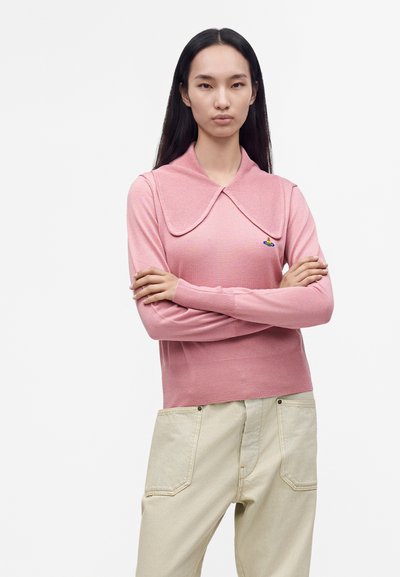 Jeune femme aux longs cheveux noirs portant un pull rose à grand col et un pantalon de couleur crème, debout les bras croisés.