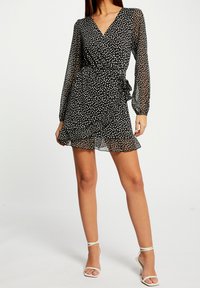 Vestido cruzado negro con lunares blancos, mangas largas transparentes y dobladillo con volantes. Estilizado con sandalias blancas de tacón para crear contraste.