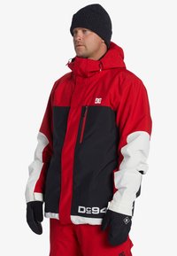 Rød, sort og hvid snowboardjakke med en lynlåslomme, justerbar hætte og logodetaljer. Sammen med sorte vanter og bukser.