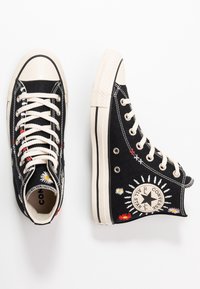 Černé plátěné vysoké tenisky s bílou gumovou podrážkou, zdobené květinovou výšivkou a béžovými šněrovadly. Ikonické logo na vnější straně kotníku.