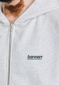 Grå hættejakke med lynlås lavet af blødt stof, med et sort og grønt "bareen" logo på venstre brystområde. Glat tekstur, afslappet design.