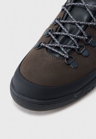 Καφέ μποτάκι πεζοπορίας με μαύρο προστατευτικό μύτης, επενδυμένη μαύρη γλώσσα και γκρι κορδόνια με σχέδιο περασμένα μέσα από μεταλλικά κουμπώματα.