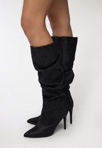 Czarne, luźne botki do kolan z ostrym noskiem, wykonane z teksturowanej tkaniny, ozdobione małymi, odblaskowymi detalami; posiadają zamek błyskawiczny.