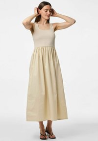 Bež maxi haljina s pripijenim gornjim dijelom i naboranom suknjom. Napravljena od glatkog materijala, s kvadratnim izrezom i tankim trakama.