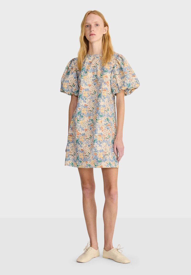 Femme portant une robe courte fleurie avec des manches bouffantes et des chaussures écrues à lacets, debout devant un fond uni.
