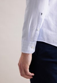 Polsino di camicia a righe azzurro chiaro con due bottoni bianchi e un emblema floreale ricamato sul lato esterno. Tessuto in cotone liscio.