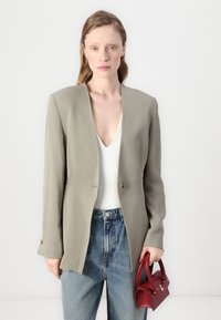 Lys olivengrønn blazer med skreddersydd fasong, enkel knappelukking og lange ermer. Kombinert med en hvit topp og blå høy midje jeans, holder en liten rød håndveske.