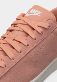 Ροζ αθλητικό παπούτσι Nike από σουέντ με ροδάκινι κορδόνια, λευκό λογότυπο Nike στη γλώσσα και λευκή καουτσούκ σόλα σε λευκό φόντο.