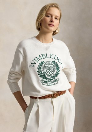Femme aux cheveux blonds courts portant un pull blanc avec le logo de Wimbledon, un pantalon blanc, une ceinture tressée marron et une seule boucle d'oreille pendante, les mains dans les poches.