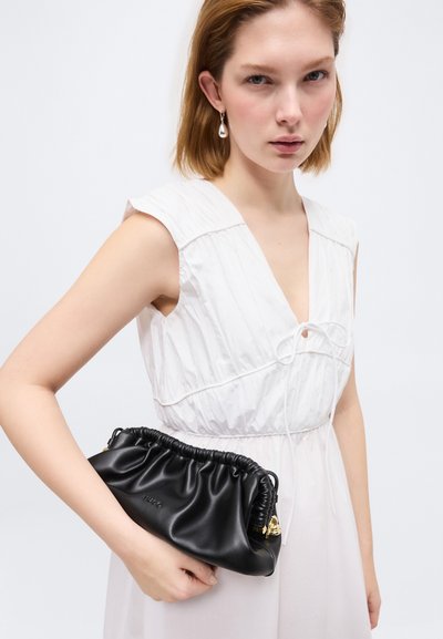 Jeune femme en robe blanche sans manches tenant une pochette en cuir noir plissé avec des accents dorés et le nom de la marque "Hugo".