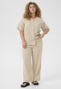 Camicia beige a maniche corte con vestibilità comoda e chiusura con bottoni, abbinata a pantaloni a gamba larga. Texture liscia; dettagli minimi; sandali con tacco scuro.