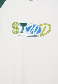 Chemise blanche avec des lettres stylisées colorées "STWD" en vert, bleu et jaune sur la poitrine, chaque lettre présentant un design unique.