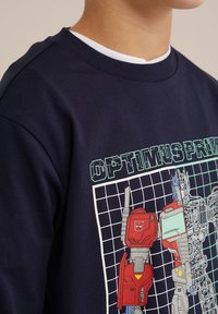 Tamsiai mėlynas medvilninis džemperis su Optimus Prime grafika raudonos ir melsvos spalvos, esantis ant tinklinio fono. Apvali iškirptė ir lygi tekstūra.