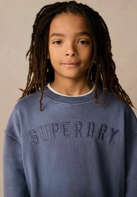 Felpa blu con "SUPERDRY" ricamato in blu navy. Tessuto morbido, scollo rotondo e vestibilità comoda con dettagli di cucitura visibili.