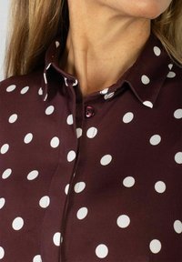 Bordowa bluzka w białe groszki, z ostrym kołnierzem i różowym guzikiem. Tkanina wydaje się gładka z lekkim połyskiem.