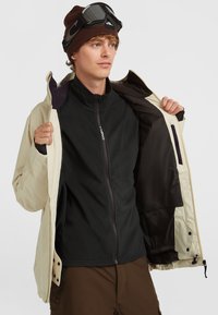 Beige buitenjacket met zwarte fleece binnenvoering, rits aan de voorkant en verstelbare manchetten. Draag open over een zwart fleece shirt, met een bruine beanie en een paar goggles.