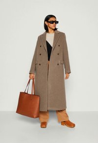 Mulher vestindo um longo casaco castanho, calças beges, tamancos castanhos, óculos de sol pretos, segurando uma grande bolsa de tote de couro castanho.