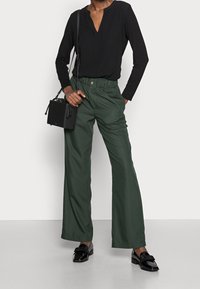 Un haut noir à manches longues avec un col en V, associé à un pantalon vert à taille haute et à jambes larges. Les accessoires comprennent un petit sac à main noir et des chaussures noires.