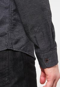 Mørkegrå langærmet skjorte med tekstureret stof, der har en knaplukket manchet og en lomme på siden. Geometrisk stikningsdetalje synlig.