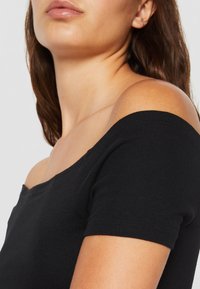 Gros plan d'une personne portant un haut noir décolleté montrant son épaule, son cou et ses lèvres sur un fond uni.