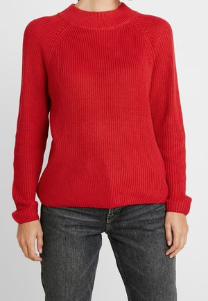 Personne portant un pull en maille côtelée rouge à manches longues et un jean noir délavé, debout devant un fond uni.