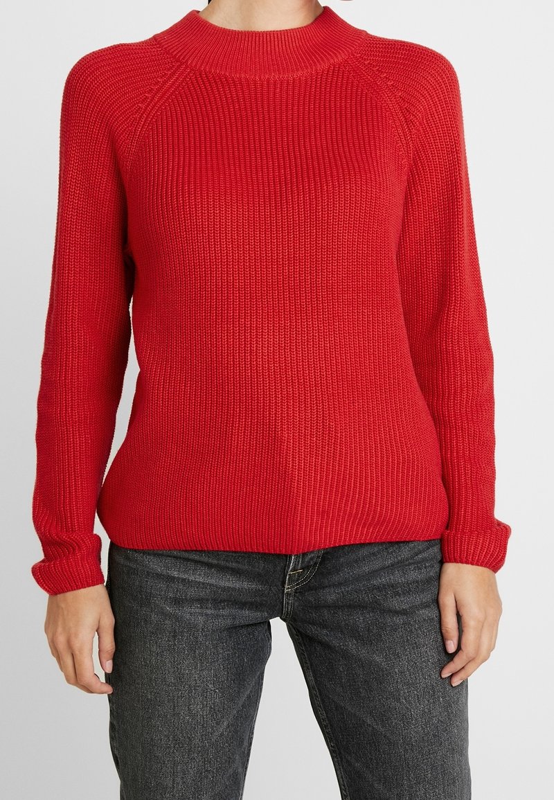 Personne portant un pull en maille côtelée rouge à manches longues et un jean noir délavé, debout devant un fond uni.
