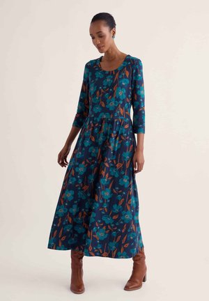Femme portant une longue robe bleu marine avec des motifs floraux bleus et des motifs de feuilles marron, manches trois-quarts, associée à des bottines marron.