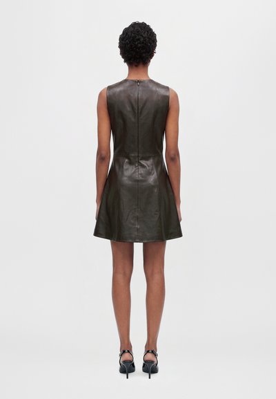 Un mannequin vu de dos porte une mini-robe en cuir sombre sans manches et des talons noirs sur un fond blanc uni.